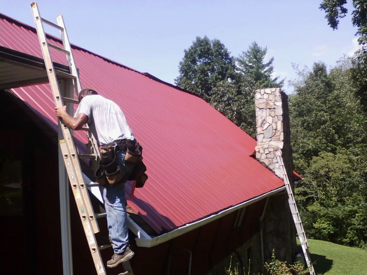 Roofer on a ladder beside a red metal roof and stone chimney — Soffit & Fascia Repair by Roofing Brothers in Roxbury Crossing